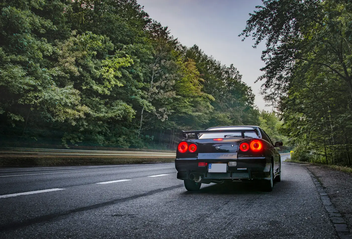 A dark Nissan Skyline R34 GT-T seen from behind on a road through trees