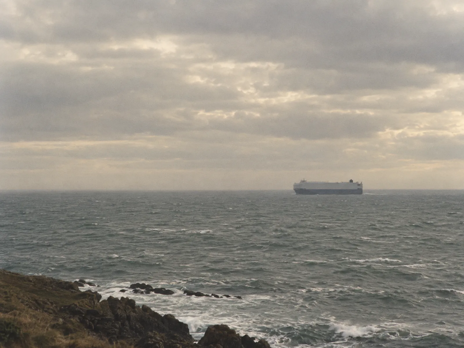 Illustration of a car-carrier ship far out at sea under an overcast sky, seen from a rocky headland